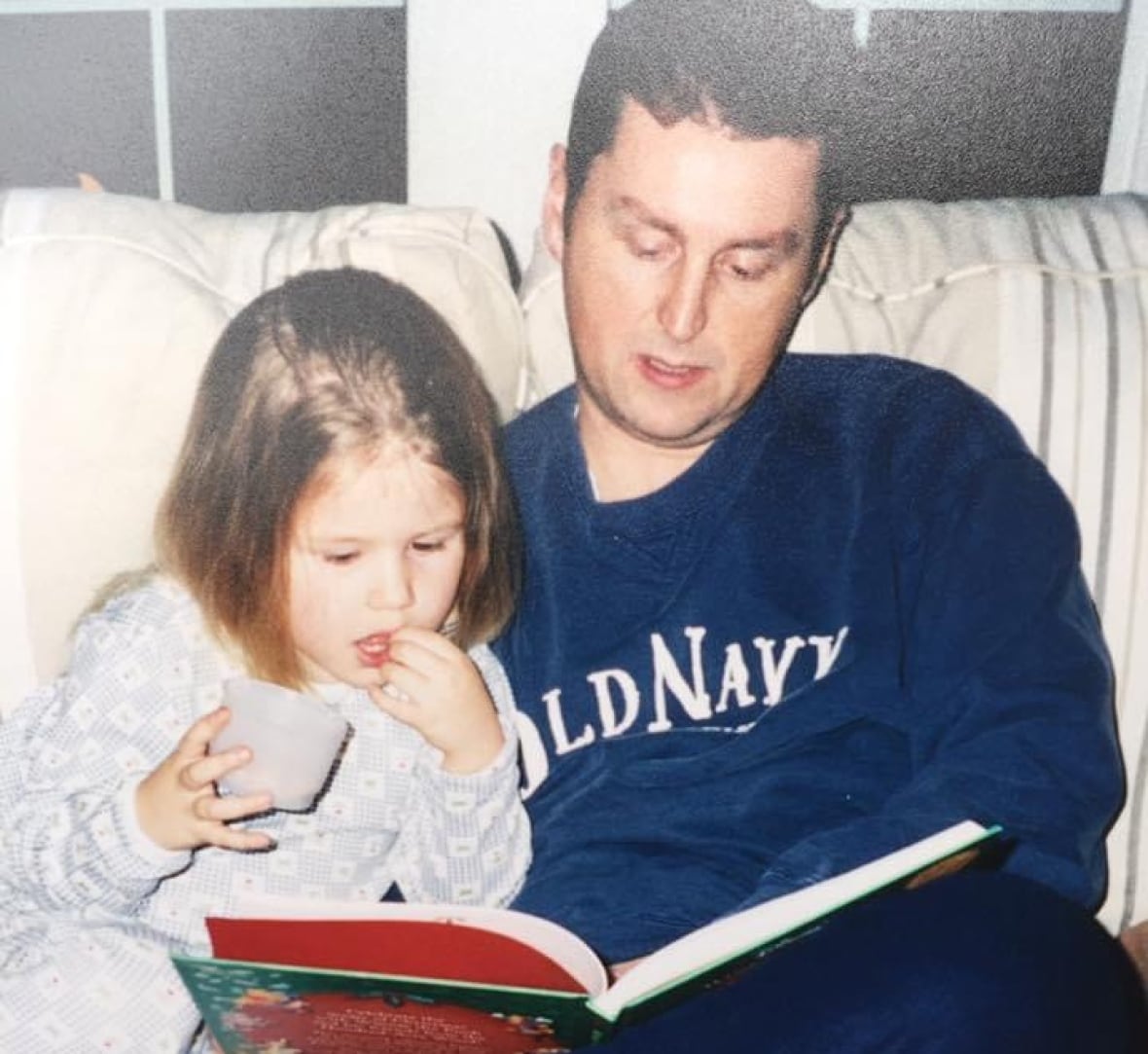 A young child with a bob sits on a couch looking at a picture book with a man in an Old Navy sweatshirt. 