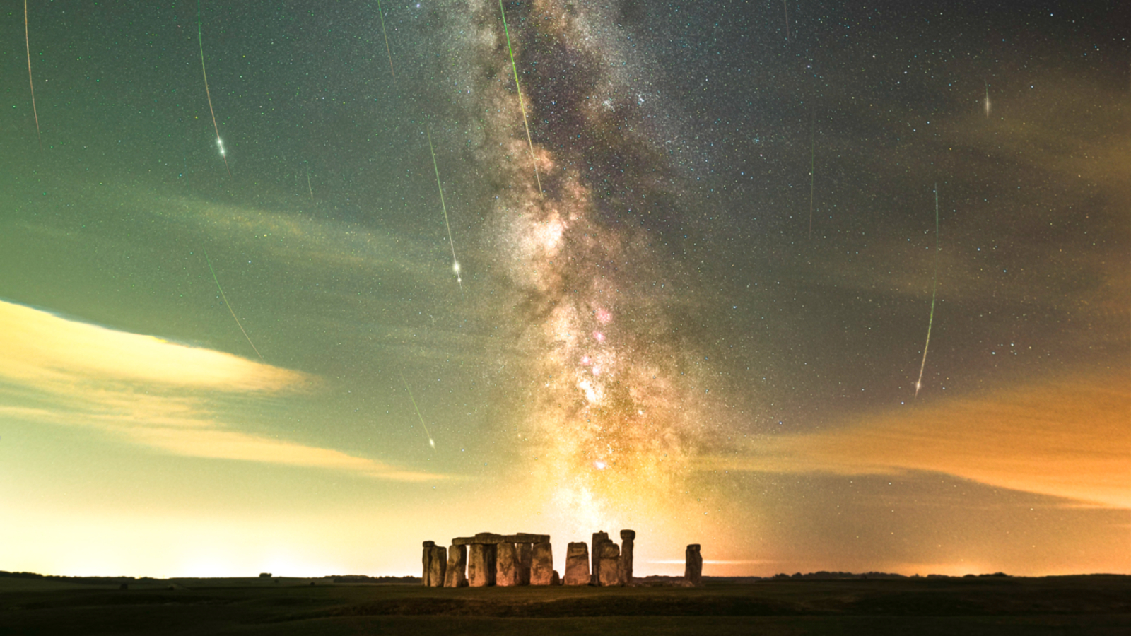 The ancient monument Stonehenge is pictured at night with the Milky Way glowing in a band tumbling towards the horizon. Pereid meteor trails can be seen arcing towards the historic site in a night sky dotted with stars.