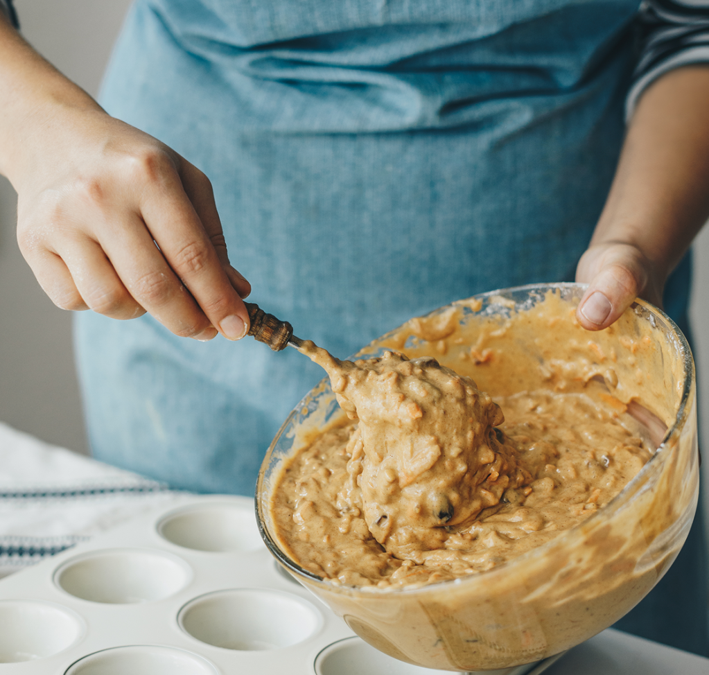 Hands prepare to spoon thick batter from a glass bowl into a waiting muffin tin, ready for baking.