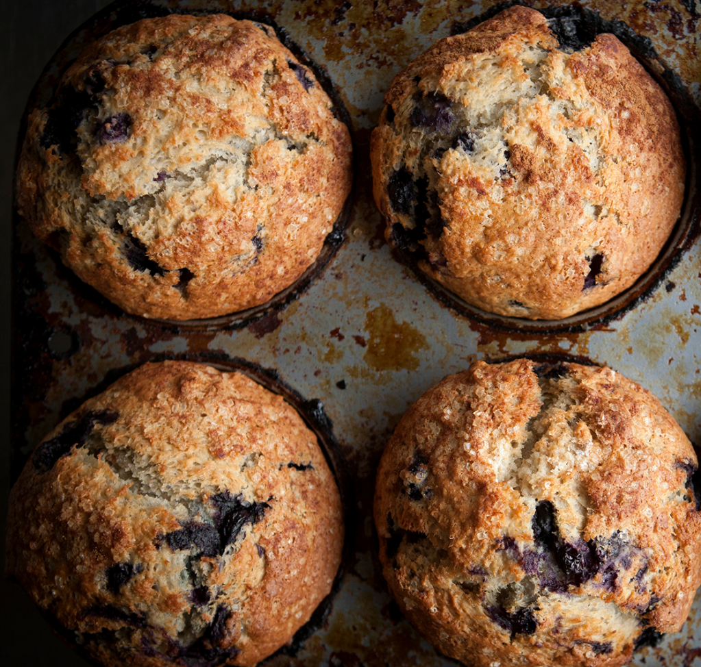 Four golden-brown blueberry muffins rest in a well-used muffin tin, fresh from the oven.