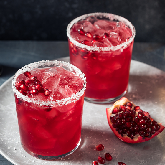 Two glasses of bright red pomegranate cocktails with sugared rims and pomegranate seeds sit on a tray beside a fresh pomegranate half.