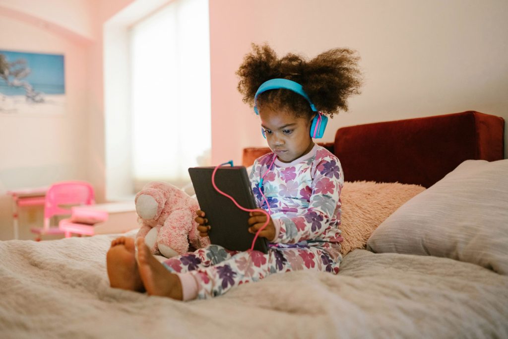 A little girl listens to something on an iPad with headphones while sitting on a bed in her room.