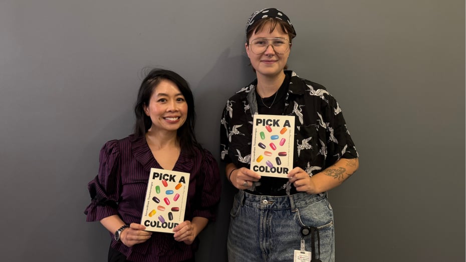 A woman and a person hold the same book and smile in front of a grey wall.