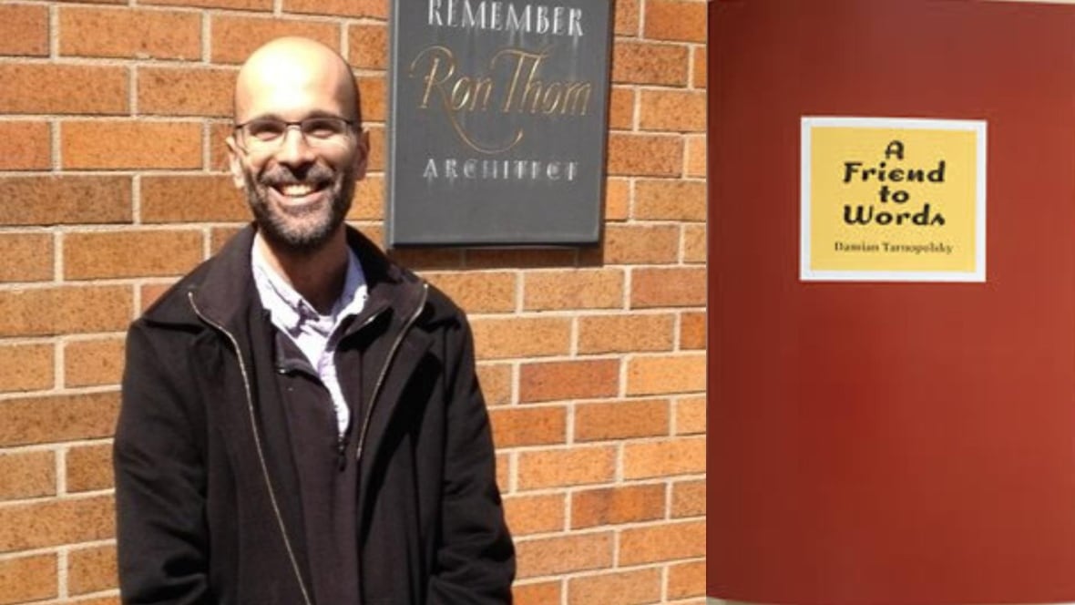 A smiling man with a beard wearing glasses and smiling at the camera while standing in front of a brick wall and the red book cover