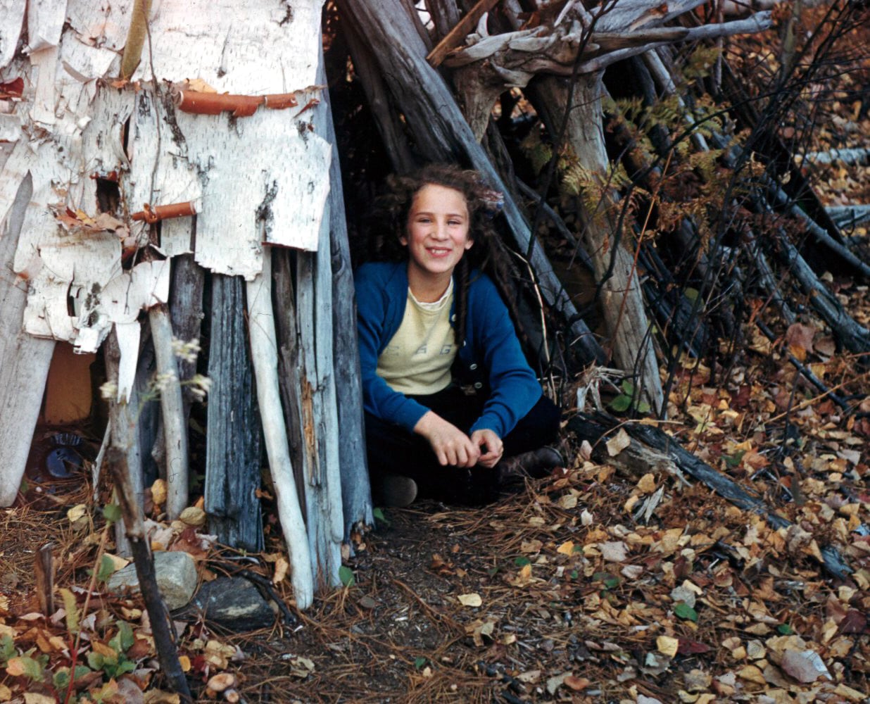 A young girl sits at the door of a wooden hut, out in the woods