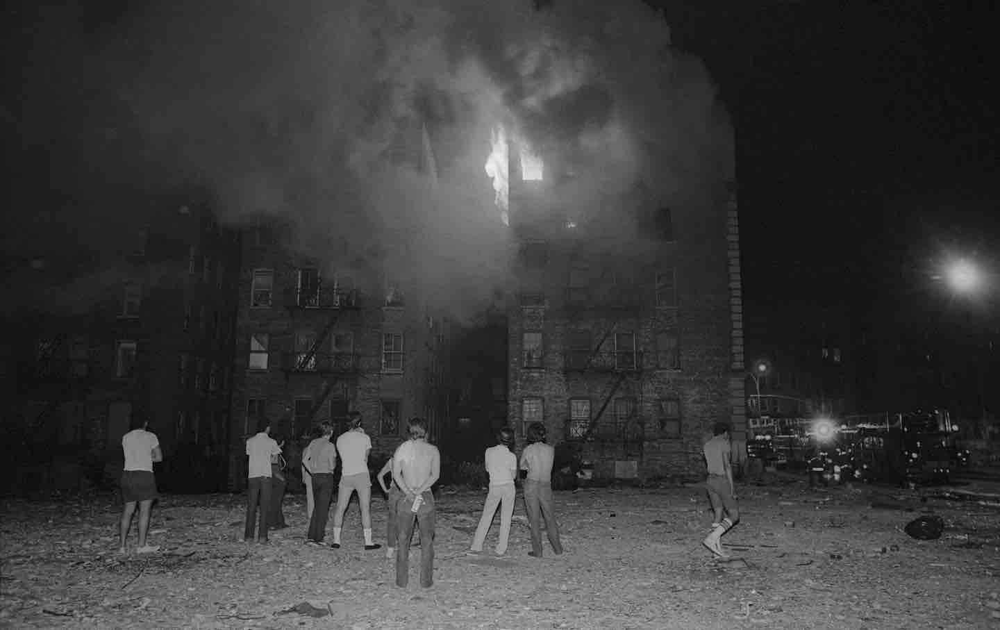 Nighttime view of people in a vacant lot as they watch a fire burning on the top floors of an apartment building in the Bronx, New York, 1983.